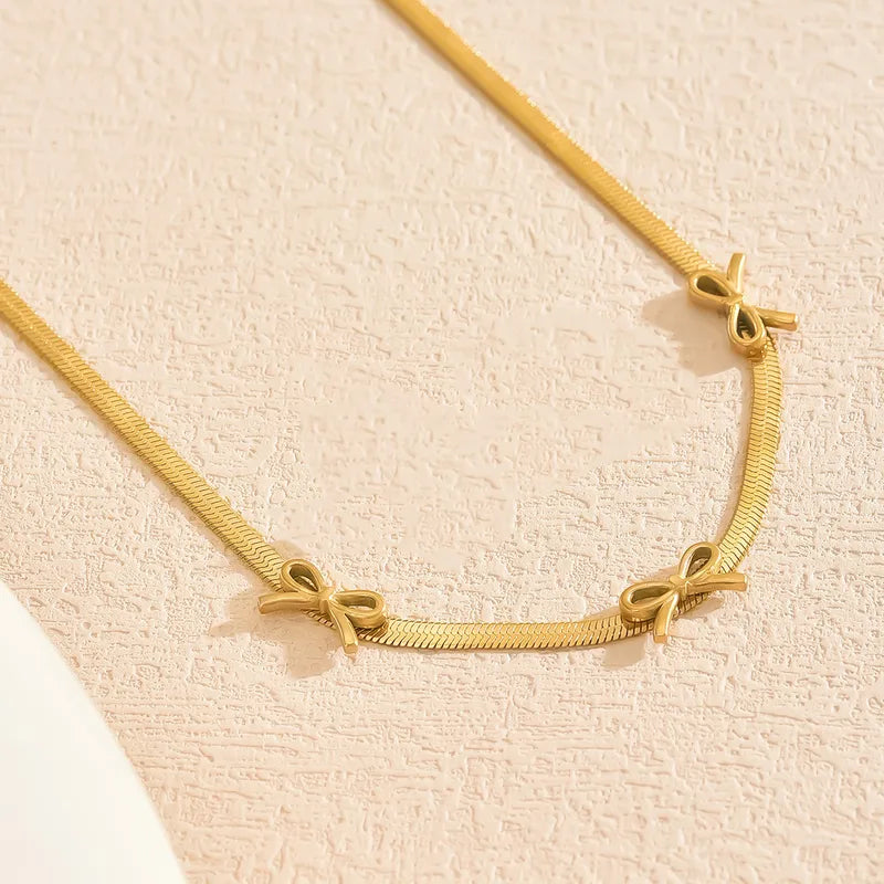 H13 - Collar planchado con dijes de moñitos. Color dorado. Acero inoxidable.